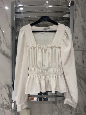 Vit blus från Chiquelle - Vit blus från Chiquelle med smockdetaljer över byst och midja. Blusen har långa puffärmar och volang nedtill. 