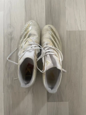 Vita Adidas fotbollsskor med guld - Säljer ett par vita Adidas fotbollsskor med guldiga detaljer och klassiska tre ränder på sidan. Skorna har snörning, låg profil och dobbar för spel på gräs. Tillverkade i syntetmaterial för lätt vikt och bra bollkontroll. Perfekta för dig som vill sticka ut på planen.
