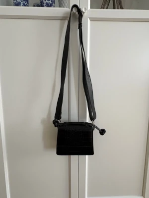 Svart axelväska med krokopräglat mönster - Snygg svart axelväska i konstläder från Carin Wester med krokopräglat mönster och justerbar axelrem. Väskan har en fyrkantig form och lockstängning. Perfekt storlek för det viktigaste och ger en trendig touch till din outfit.