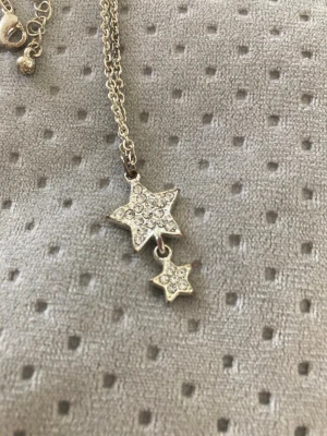 Silverfärgat halsband med stjärnhängen - Snyggt halsband i silverfärg med två stjärnformade hängen, båda dekorerade med små glittrande stenar. Kedjan är tunn och har justerbar längd med ett litet klot vid låset. Perfekt för dig som gillar detaljer och stjärntema. Silverfärgen har kommit av vissa delar , men fortfarande mycket kärlek att ge till någon.