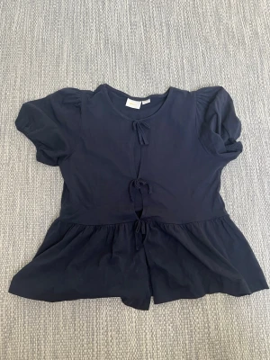 Mörkblå blus med knyt från Gina Tricot - Supersöt mörkblå blus från Gina Tricot med korta puffärmar och tre knyt framtill. Blusen har en lätt utsvängd passform och är gjord i mjukt tyg.