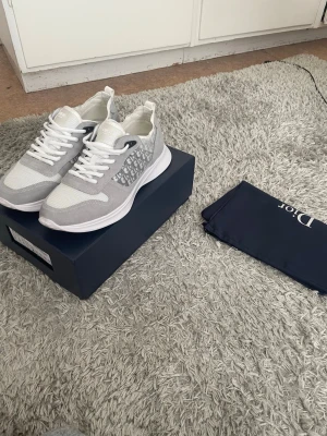 Gråvita Dior sneakers i mesh - Snygga sneakers från Dior i grått och vitt med mesh- och mockadetaljer. Skorna har vita snören och en chunky sula som ger en modern vibe. Perfekta för dig som gillar exklusiva märken och stilren design.