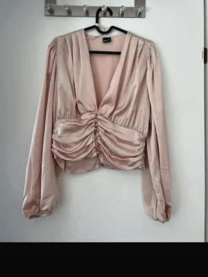 Ljusrosa satinblus från Gina Tricot - Trendig ljusrosa blus i satin från Gina Tricot med djup v-ringning och snygg rynkning framtill. Liten i storleken 