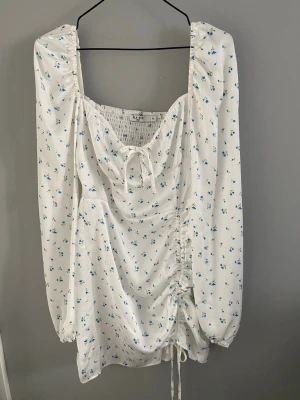 Jättefin adooreliknande kortklänning från NA-KD x Sanna Jörnvik - Söt vit kortklänning från NA-KD med små blommor, långa puffärmar och snörning framtill. Klänningen går att dra upp på ena sidan, har en sidenkänsla och en adooreliknande passform som känns både snygg och trendig. Perfekt för sommarens alla tillfällen! Använd endast en gång - mycket fint skick!