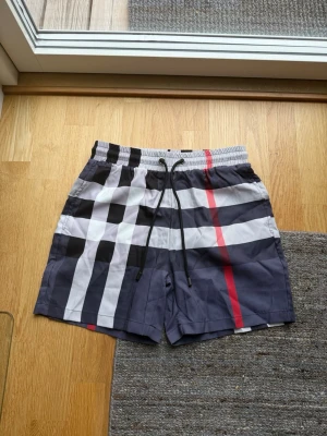 Rutig badshorts från Burberry - Säljer ett par klassiska badshorts ”som även kan användas som vanliga shorts” från Burberry med ikoniskt rutmönster i svart, vitt och rött. Shortsen har snörning i midjan och är tillverkade i ett snabbtorkande syntetmaterial. Perfekta för sommaren och strandhänget.