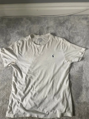 Vit t-shirt från Polo Ralph Lauren - Klassisk vit t-shirt från Polo Ralph Lauren med rund halsringning och den ikoniska broderade loggan på bröstet. Tillverkad i mjuk bomull för en skön känsla och enkel att matcha till olika outfits.