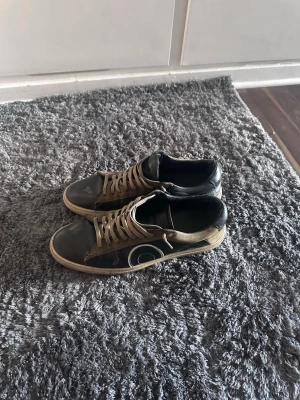 Svarta sneakers med beige detaljer från oliwer cabell - Snygga sneakers i svart och beige med vita snören och en unik cirkeldesign på sidan. Skorna har låg profil och en beige sula. Materialet ser ut att vara en mix av läder och textil, vilket ger en cool och urban vibe.