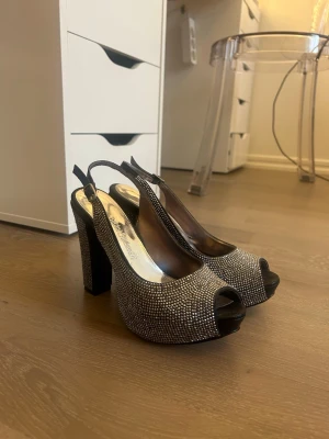 Glittriga peep toe pumps från Bianco - Säljer ett par svarta pumps med öppen tå och slingback-rem. Skorna har höga blockklackar och är täckta med små glittrande stenar som ger en lyxig känsla. Perfekta för dig som vill sticka ut och glänsa lite extra.