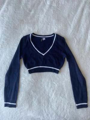 Mörkblå stickad magtröja - Mörkblå stickad magtröja med vita ränder vid v-ringad hals, midja och ärmslut. Långärmad modell som ger en sportig och fräsch look. 