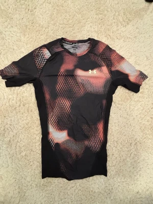 Under Armour kompressionströja mönstrad - Säljer en tight kompressionströja från Under Armour med coolt ormskinnsmönster i svart, rött och orange. Tröjan har korta ärmar, rund hals och är tillverkad i ett tekniskt funktionsmaterial som andas. Perfekt för träning eller gympass.