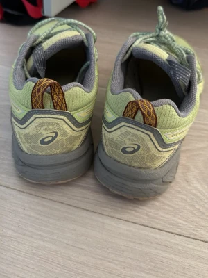 Ljusgröna Asics sneakers - Säljer ett par ljusgröna sneakers från Asics med grå detaljer och mönstrade dragflikar i brunt och orange. Skorna har mesh-ovandel för bra ventilation, robust sula och klassisk snörning. Perfekta för dig som gillar sportig stil och bekväma skor.