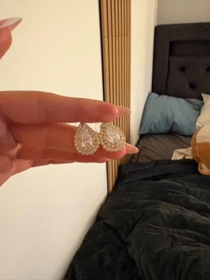Glittriga droppformade örhängen - Säljer ett par droppformade örhängen i guldton med gnistrande stenar och pärldetaljer. Örhängena har en elegant och lyxig look med flera små pärlor runt en större sten i mitten. Perfekt för dig som gillar att sticka ut med accessoarer.