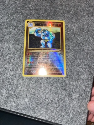 Machoke holografiskt Pokémonkort - Machoke holografiskt samlarkort från Pokémon med gul ram och glansig yta. Kortet har klassisk blå baksida med Pokéball och texten Pokémon. Perfekt för dig som gillar att samla eller spela Pokémonkort.