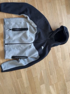 Nike tech fleece - Nike hoodie i svart och grå med hel dragkedja framtill och två svarta dragkedjeförsedda fickor. Hoodien har en sportig look med huva och är tillverkad i mjukt material, perfekt för chill eller träning.