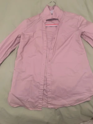 Ljusrosa skjorta med knappar - Snygg ljusrosa skjorta med klassisk krage och knäppning framtill. Skjortan har lång ärm och är tillverkad i mjuk bomull, perfekt för en clean och stilren look. Passar till både jeans och kostymbyxor.