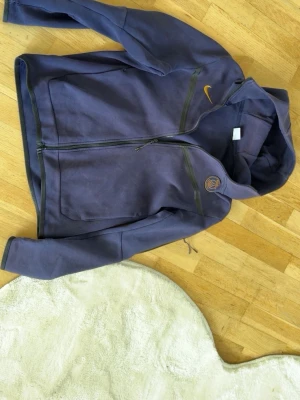 Lila Nike hoodie med dragkedja - Snygg lila hoodie från Nike med dragkedja framtill och två stora fickor. Har huva och diskret Nike-logga i gult på bröstet samt ett tryck på andra sidan. Tillverkad i mjukt bomullsmaterial som känns skönt mot huden.