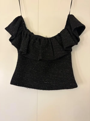 Glittrig svart off shoulder topp - Snygg svart topp med glittertrådar och bred volang runt axlarna. Off shoulder model och figurnär passform.