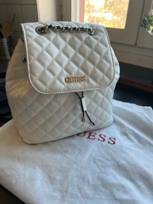 Vit quiltad ryggsäck från Guess - Snygg vit ryggsäck från Guess i quiltat i vegansk läder  guldfärgade detaljer och kedjehandtag. Väskan har svarta justerbara axelband och dragskostängning. Perfekt för dig som vill ha en trendig och stilren accessoar till vardagslooken.