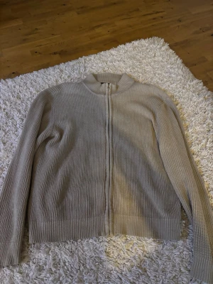 Beige ribbad ziptröja från 157 - Säljer en beige ribbstickad ziptröja från 157 med hög krage och hel dragkedja framtill. Tröjan har långa ärmar och är perfekt för lager på lager. Enkel och stilren design som passar till det mesta.