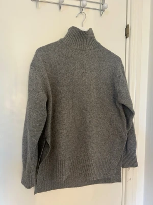 Grå polotröja från H&M - Mysig grå stickad polotröja från H&M med hög krage och ribbade muddar. Tröjan har en avslappnad passform och är perfekt för kyliga dagar. Materialet är mjukt och värmande, vilket gör den superbekväm att bära.