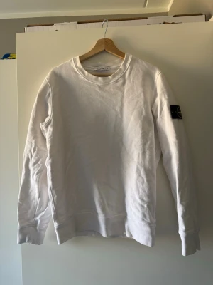 Vit sweatshirt från Stone Island - Säljer en clean vit sweatshirt från Stone Island med den ikoniska svarta patchen med gult och grönt märke på vänster ärm. Tröjan har rund hals, långa ärmar och är gjord i mjukt bomullsmaterial.  Bra skick. Storlek S. 