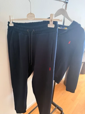 Ralph Lauren - Snyggt mörkblått set från Ralph Lauren med matchande byxor och zip-hoodie. Båda plaggen har det klassiska röda Polo-logot broderat på framsidan. Setet har en sportig look med dragsko i midjan och fickor på både byxor och hoodie. Perfekt för en avslappnad stil.