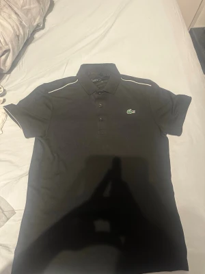 Svart pikétröja från Lacoste - Klassisk svart pikétröja från Lacoste med vit piping längs axlarna och den ikoniska gröna krokodilloggan på bröstet. Tröjan har korta ärmar, krage och knappar framtill. Perfekt för en clean och sportig look.