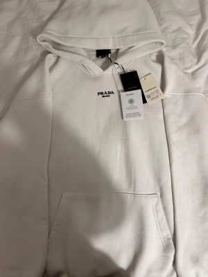 Vit hoodie från Prada - Säljer en clean vit hoodie från Prada med svart logga broderad på bröstet. Hoodien har stor huva, känguruficka framtill och ribbade muddar. Tillverkad i mjuk bomull för skön känsla. Perfekt för dig som gillar exklusiva streetwear-plagg.