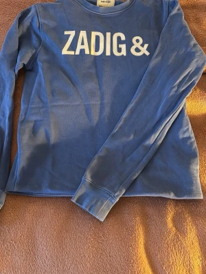 Blå långärmad tröja Zadig & Voltaire - Snygg blå långärmad tröja från Zadig & Voltaire med vit text på både fram- och baksidan. Tröjan har rund halsringning och är tillverkad i mjukt bomullsmaterial. Perfekt för en avslappnad och trendig look. Använt få gånger.