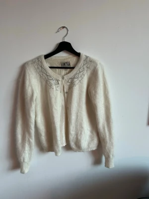 Vit fluffig kofta med paljetter - Säljer en vit, fluffig kofta med rund hals och dekorativa silverpaljetter och mönster runt halsringningen. Koftan har långa ärmar och knäpps framtill med små knappar. Perfekt för dig som gillar en mjuk och glittrig stil. Det är 50% angora ull och 50% nylon så den är SUPERMJUK 🌸☀️ silke inuti 