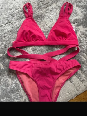 Rosa bikini med dubbla axelband - Snygg rosa bikini från Bodytune med dubbla axelband och trendiga banddetaljer runt midjan. Överdelen har triangelformade kupor och justerbara band, medan underdelen har en klassisk passform. Perfekt för strandhäng eller poolparty!