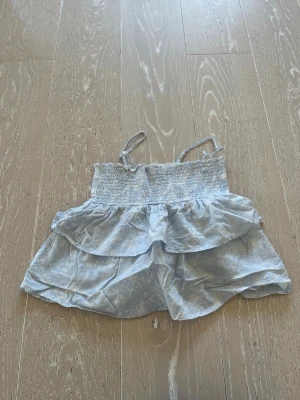 Ljusblå volangtop från Gina Tricot - Supersöt ljusblå topp från Gina Tricot med smala axelband och dubbla volanger. Toppen har smock upptill och ett diskret blommigt mönster. Perfekt för varma dagar och ger en fräsch sommarkänsla.