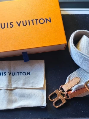 Louis Vuitton axelrem beige - Säljer en beige axelrem från Louis Vuitton med justerbar längd och detaljer i guldmetall och läder. Axelremmen har diskret Louis Vuitton-logga invävd i tyget och kommer med originalförpackning och dustbag. Kom med min High Rise Bumbag från Vestiaire Collective.