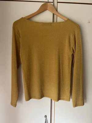 Gul glittrig långärmad topp - Snygg gul långärmad topp med glittrigt material och enkel, rak passform. Perfekt för dig som vill sticka ut lite extra. Toppen har en rund halsringning och är tillverkad i ett stretchigt tyg som känns bekvämt mot huden.