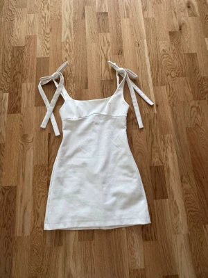 Vit miniklänning med knytband - Snygg vit miniklänning med smickrande passform och knytband på axlarna. Klänningen har en rak urringning och är ärmlös, vilket ger en fräsch och somrig look. Perfekt till student eller sommarens alla tillfällen. Materialet känns mjukt och lätt.