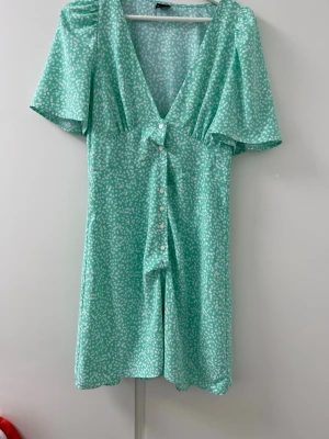 Grön blommig kortklänning med knappar - Söt grön kortklänning med vitt blommönster och v-ringning. Klänningen har korta, vida ärmar och knappar framtill. Perfekt för varma dagar och midsommar.  Klänningen har en avslappnad luftig passform.