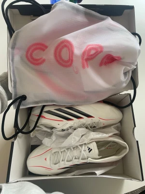 Adidas Copa vita fotbollsskor - Säljer ett par helt nya vita Adidas Copa fotbollsskor med svarta och röda detaljer. Skorna har snörning, låg profil och är tillverkade i syntetmaterial med Fusionskin-teknologi. Medföljer en vit påse med röd Copa-logga. Perfekta för dig som vill ha stilrena och lätta skor på planen.