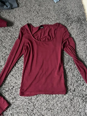 Vinröd långärmad topp Gina Tricot - Snygg vinröd långärmad topp från Gina Tricot med rund, djup urringning. Tillverkad i mjukt och stretchigt material som sitter skönt på kroppen. Perfekt att matcha med jeans eller kjol för en trendig look.