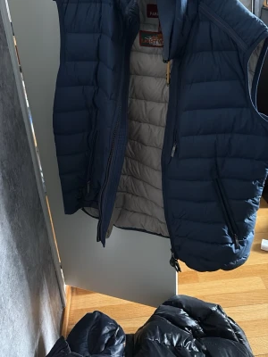Blå dunväst från Parajumpers - Snygg blå dunväst från Parajumpers med quiltad design och beige insida. Västen har hög krage, dragkedja framtill och sidofickor med dragkedja. Märkt med 'Super Lightweight' vilket gör den perfekt för lager-på-lager och chill stil. Litet hål på baksidan