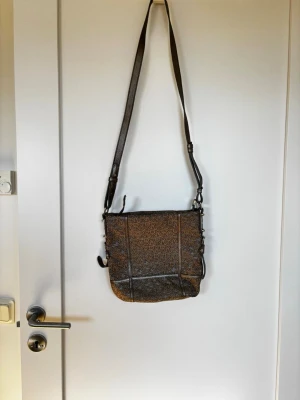 Brun axelväska från DKNY - Snygg brun axelväska från DKNY med justerbar axelrem och klassiskt DKNY-mönster över hela väskan. Väskan har detaljer i mörkbrunt konstläder och en praktisk form som rymmer det mesta du behöver. Perfekt för dig som gillar stilrena accessoarer med logotypmönster. Använd bara en gång. 