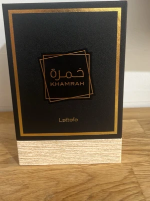 Lattafa Khamrah Eau de Parfum 100ml - Lattafa Khamrah Eau de Parfum kommer i en lyxig svart och guld förpackning. Flaskan är fyrkantig med ett elegant mönster i glaset och bär en djup bärnstensfärgad vätska. Märket Lattafa och namnet Khamrah syns tydligt på både låda och flaska. Volymen är 100 ml.