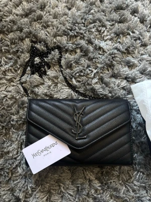 Svart YSL handväska med kedjerem pris kan diskuteras! - Säljer en svart handväska från Yves Saint Laurent med klassisk YSL-logga framtill. Väskan har quiltat mönster, kedjerem i svart metall och kuvertformad design. Kommer med dustbag och originalbox. Perfekt för dig som vill ha en lyxig accessoar.
