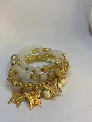 Vita och guldiga armband med fjärilar - Snygga armband i vitt och guld med pärlor och detaljerade fjärilsberlocker i guld. Armbanden är elastiska och består av olika former av pärlor, både runda och facetterade, som ger en lyxig och trendig känsla. Ej använd utan kommer från min small business.