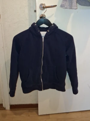 Mörkblå zip hoodie med huva - En enkel och stilren mörkblå hoodie med dragkedja framtill och två fickor. Hoodien har en klassisk huva med snörning och ribbade muddar vid ärmar och nederkant. Perfekt för en avslappnad look och passar till det mesta.