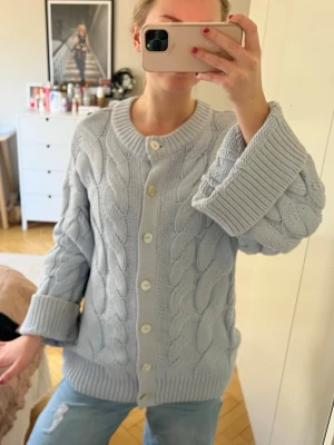 Ljusblå kabelstickad kofta - Mysig ljusblå kofta med kabelstickat mönster och stora vita knappar framtill. Koftan har rund halsringning, ribbade muddar och en oversized passform. Perfekt för lager på lager och chill dagar.