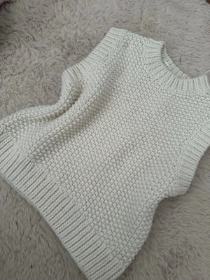 Vit stickad väst med grov struktur - Säljer en chunky stickad väst i offwhite med bred ribbad kant vid hals, ärmhål och nederkant. Västen har en grovstickad struktur som ger en cool och trendig look. Perfekt att styla över en skjorta eller t-shirt för en avslappnad vibe.