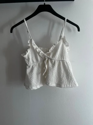 Vit linnetopp med volang och knyt - Söt vit linnetopp med tunna axelband, volangdetaljer vid ringningen och knyt framtill. Toppen har en lätt rynkad struktur och är croppad i modellen, perfekt för varma dagar.