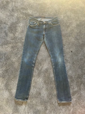 Blå skinny jeans från Nudie Jeans - Säljer ett par klassiska blå skinny jeans från Nudie Jeans med fem fickor och slitna detaljer. Jeansen har en smal passform och är tillverkade i denim med snygga slitningar på knän och bakfickor. Perfekta för en avslappnad och trendig look.