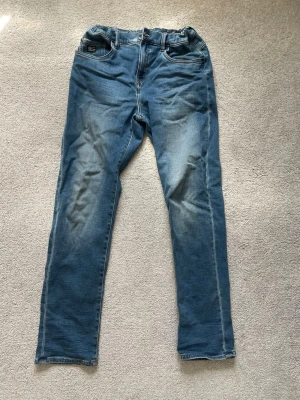 Blå raka jeans med slitningar - Säljer ett par blå jeans med raka ben och snygga slitningar på framsidan. Jeansen har klassisk femficksdesign och kontrastsömmar. Baksidan har två stora fickor. Perfekta för en avslappnad och trendig look.