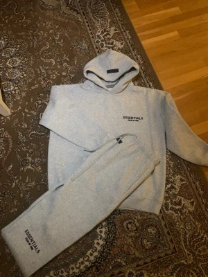 Essentials tracksuit - Mysigt loungewear-set i ljusgrått från Essentials Fear of God. Setet består av en hoodie med huva och magficka samt matchande mjukisbyxor med resår i midjan. Båda plaggen har tryckt logga på bröstet respektive benet. Perfekt för chill dagar hemma.